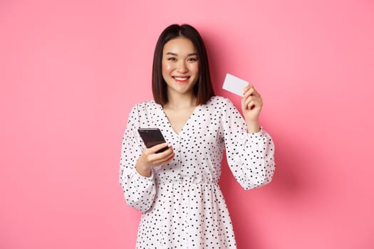 Attractive young asian woman order online, holding credit card and mobile phone, making internet purchase, standing happy over pink background.