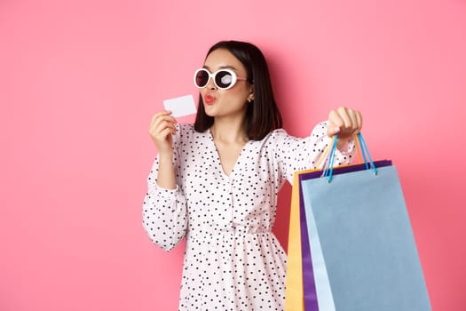 Beautiful asian woman in sunglasses going shopping, holding bags and kissing credit card, standing over pink background.