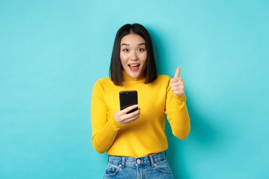 E-commerce and online shopping concept. Excited and amazed asian woman showing thumbs up after using smartphone app, recommend device, standing over blue background.