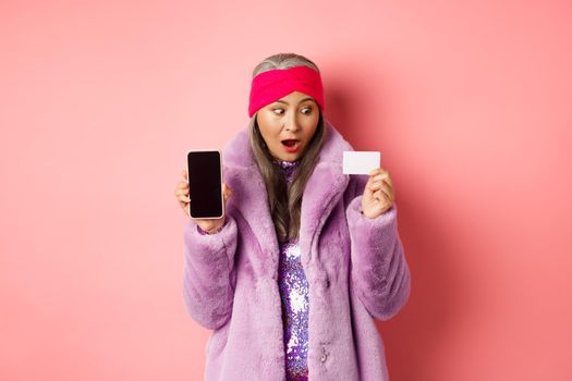 Special promotion. Excited asian senior woman showing blank smartphone screen and plastic credit card, checking out online offer, pink background.
