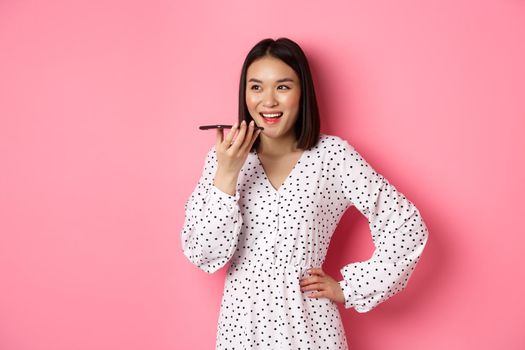 Beautiful korean woman talking on speakerphone, recording voice message and smiling happy, standing over pink background.