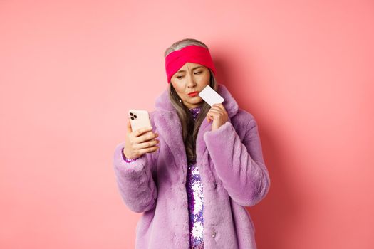 Online shopping and fashion concept. Stylish asian lady in purple faux fur coat looking indecisive at smartphone screen while holding plastic credit card, pink background.