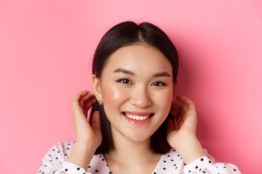 Beauty and skin care concept. Close-up of adorable smiling asian woman tuck hair behind ears, blushing and gazing at camera, standing over pink background.