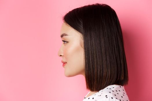 Beauty and skin care concept. Headshot profile of young beautiful asian woman with short dark hair, looking left at copy space, standing over pink background.
