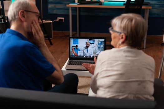Aged couple talking on video call with dentist using laptop at home. Retired people doing dentistry checkup, telemedicine appointment with specialist while sitting in living room.