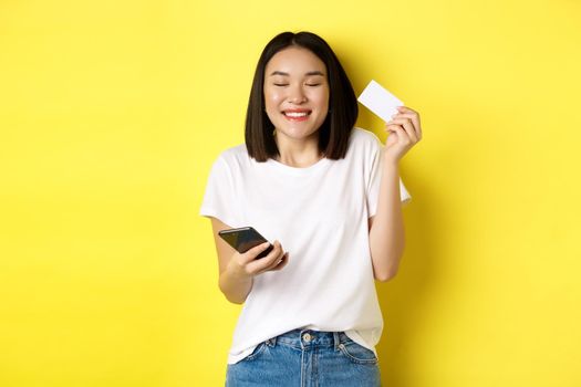E-commerce and online shopping concept. Happy asian woman looking excited, buying something in internet, holding smartphone and showing plastic credit card, yellow background.
