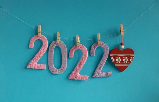 Knitted figures of 2022, made of pink and lilac threads, hang on clothespins on a blue background with a red heart. The concept of the New Year.