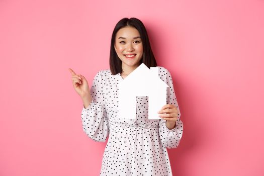Real estate. Excited asian woman showing paper house model, pointing left at copy space, standing over pink background.