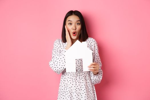 Real estate. Surprised and amazed asian woman staring at camera, saying wow and showing paper house model, standing over pink background.