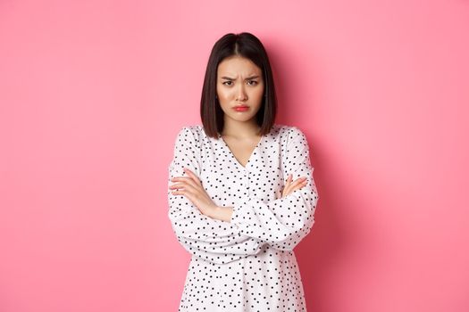 Timid and offended asian girl cross arms on chest, staring defensive and insulted at camera, standing in dress over pink background.