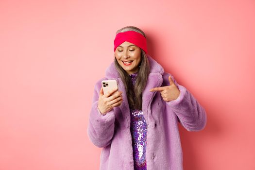 Online shopping and fashion concept. Cool asian senior woman checking out promotion on mobile phone, pointing finger at smartphone screen and smiling pleased, pink background.