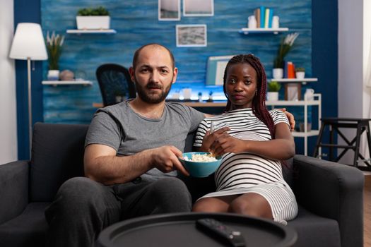 POV of interracial couple with pregnancy watching movie on TV in living room. Multi ethnic man and pregnant woman expecting baby and looking at camera while having popcorn and water