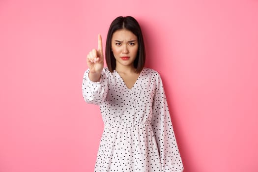 Serious and angry asian woman telling to stop, frowning and showing finger in disapproval, prohibit something, standing in dress against pink background.