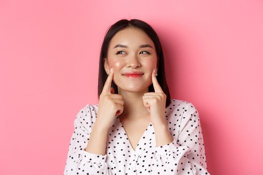 Beauty and skin care concept. Close-up of beautiful asian woman poking cheeks and smiling satisfied, standing over pink background.