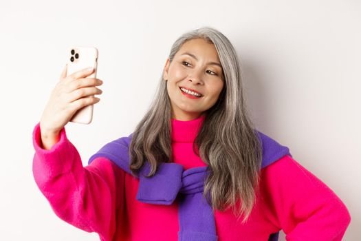 Beautiful and stylish asian senior woman taking selfie on smartphone, smiling and posing in trendy pink sweater, standing over white background.