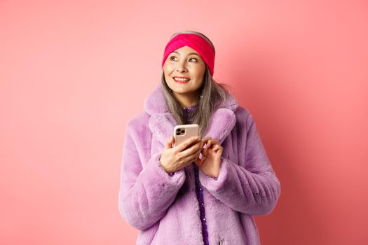 Online shopping and fashion concept. Stylish asian senior woman using smartphone, looking aside with thoughtful face, standing against pink background.