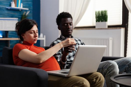 Pregnant interracial couple relaxing and using technology. Mixed race people expecting child, caucasian woman holding modern laptop and black father of baby looking at device screen