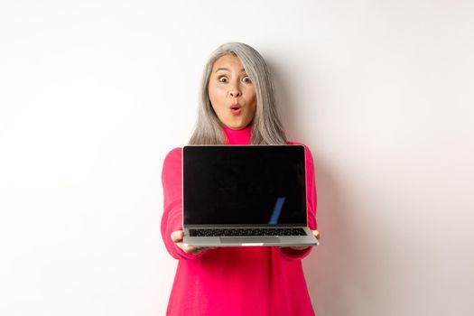 E-commerce concept. Impressed asian senior woman showing blank laptop screen and staring amazed at camera, standing over white background.