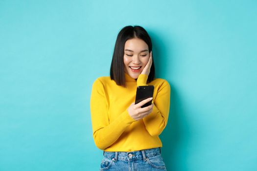 Image of happy asian woman reading message on mobile phone screen and smiling, chat in smartphone app, standing over blue background.