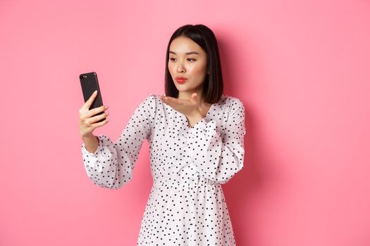 Beautiful asian girl using photo filters app and taking selfie on smartphone, posing in cute dress against pink background.