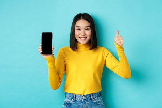 Technology and people concept. Cheerful asian girl in yellow sweater showing blank smartphone screen and thumbs up, demonstrate online offer, standing over blue background.