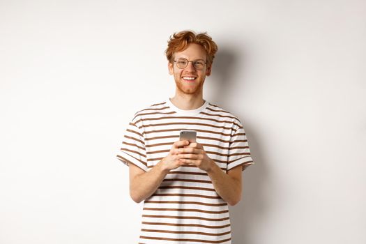 Technology and e-commerce concept. Redhead guy in glasses using mobile phone and smiling. Young man with smartphone staring happy at camera, white background.