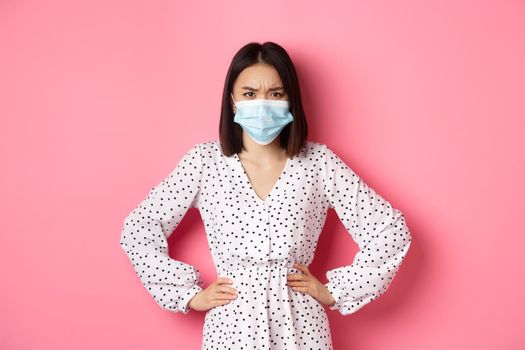 Covid-19, quarantine and lifestyle concept. Angry and disappointed korean woman in face mask, staring at camera judgemental and displeased, standing over pink background.