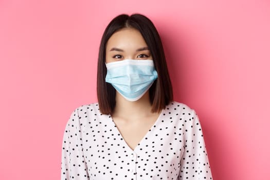 Covid-19, pandemic and lifestyle concept. Close-up of beautiful asian woman in face mask smiling with eyes, wearing protection from coronavirus in public, pink background.