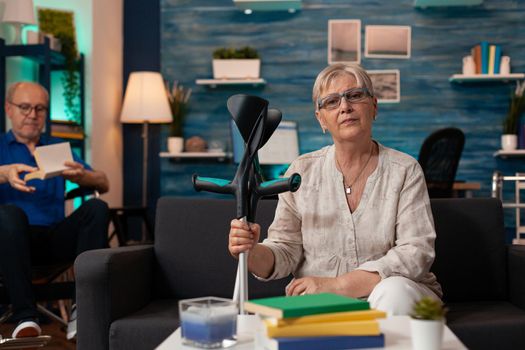 Senior woman holding crutches and looking at camera in interior space. Elder person with disability sitting on couch while retired man in wheelchair reading in living room background