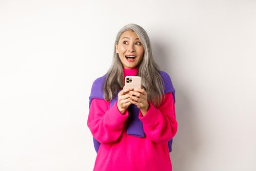 Amazed asian woman smiling and looking aside fascinated after reading promotion on smartphone, standing with mobile phone over white background.