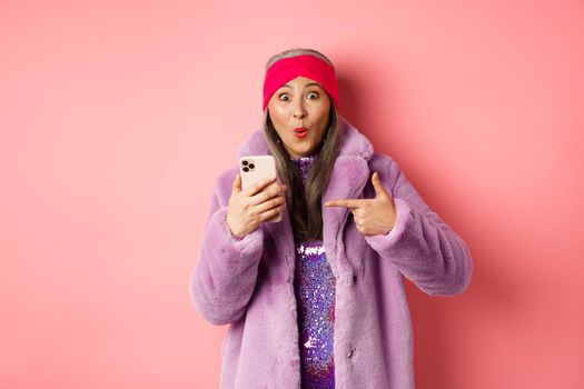 Online shopping and fashion concept. Happy asian grandmother checking out awesome promo, pointing finger at smartphone and looking amazed, pink background.