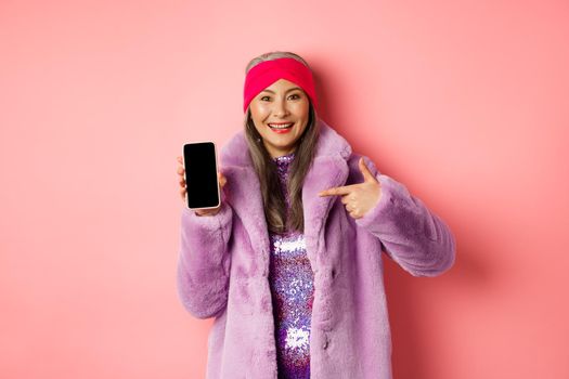 Online shopping and fashion concept. Stylish asian mature lady showing blank smartphone screen, pointing at phone and smiling, pink background.