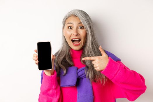 Online shopping. Beautiful asian grandmother smiling, pointing finger at smartphone blank screen, looking amazed, showing mobile application, standing over white background.