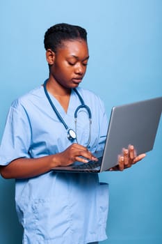 African american practitioner nurse holding laptop computer typing sickness expertise working at medical treatment in studio with blue background. Physician woman in uniform. Health care service