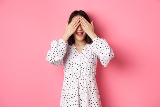 Happy romantic asian girl waiting for surprise, close eyes and smiling amazed, anticipating something, standing over pink background.