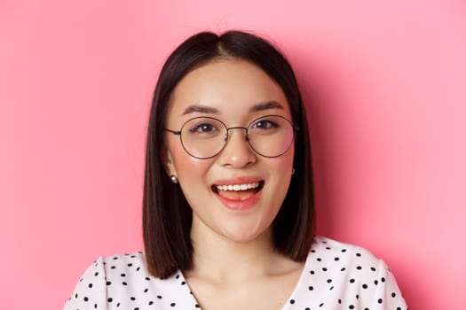 Beauty and lifestyle concept. Close-up of cute asian female model wearing trendy glasses, smiling happy at camera, standing on pink background.