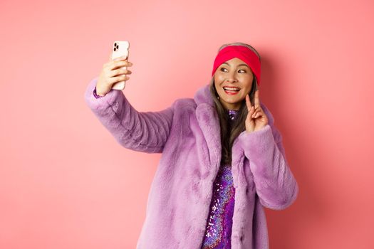 Fashion concept. Cool and cheerful asian senior woman taking selfie with peace sign, wearing trendy faux fur coat and party dress, standing over pink background.