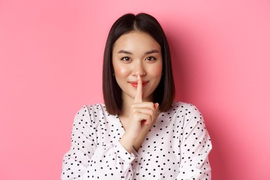 Close-up of mysterious asian woman hiding a secret, hushing and telling to keep quiet, standing over pink background.