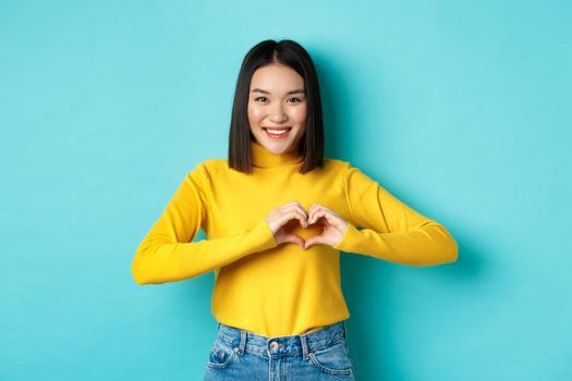 Valentines day and romance concept. Beautiful asian woman show I love you, heart gesture and smiling, standing against blue background.