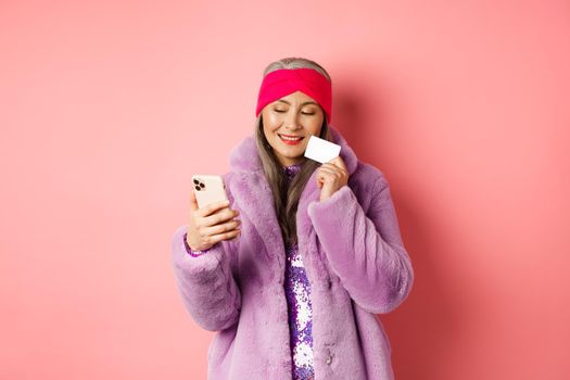 Online shopping and fashion concept. Stylish asian grandmother making purchase in internet with smartphone and plastic credit card, pink background.