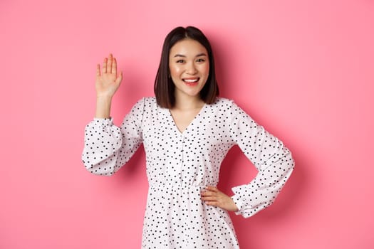 Beautiful asian woman in dress saying hello, waving hand to greet and say hi, smiling friendly at camera, standing over pink background.