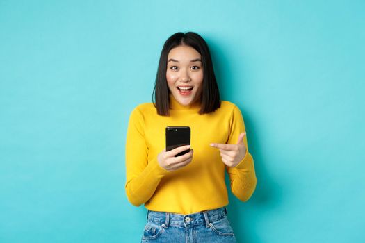 E-commerce and online shopping concept. Surprised asian woman demonstrate smartphone app, internet discounts, pointing finger at mobile phone, blue background.