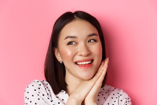 Beauty and skin care concept. Headshot of adorable and dreamy asian woman looking left, smiling and imaging, standing against pink background.