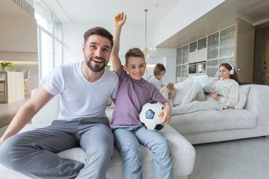Joyful father and son watch soccer at TV holding ball at home, family relaxation concept