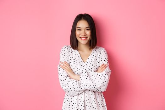 Pretty young asian woman in spring dress smiling, cross arms on chest and looking confident, standing over pink background.