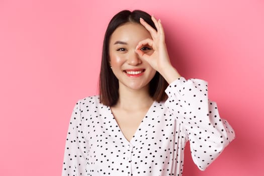 Beauty and lifestyle concept. Close-up of cute smiling asian girl showing okay sign on eye, standing over pink background in dress.