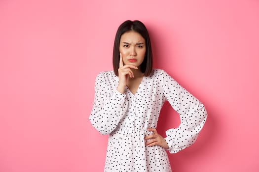 Concerned asian woman staring at camera suspicious, having doubts, thinking about something with serious face, standing against pink background.