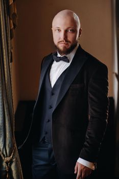 Portrait of the groom in a suit and bow tie, standing in the interior.