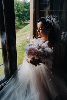 A bride in a wedding dress and a bouquet sits at an open old window and looks.