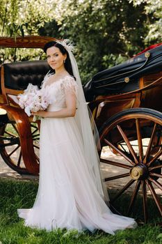 A stylish bride with a bouquet stands near a carriage in nature in retro style.